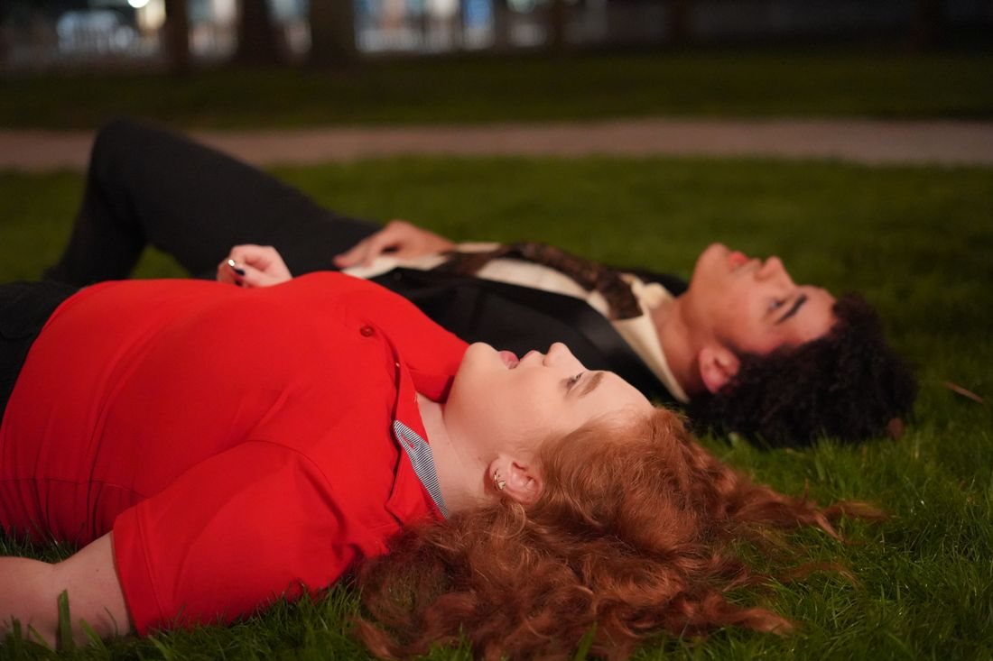 The two lead actors lie on the grass at night, looking up