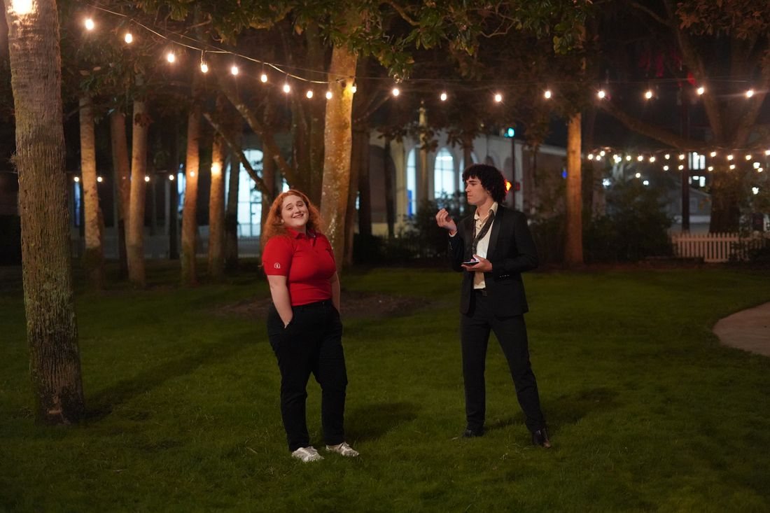 The two lead actors stand on a lawn under string lights at night