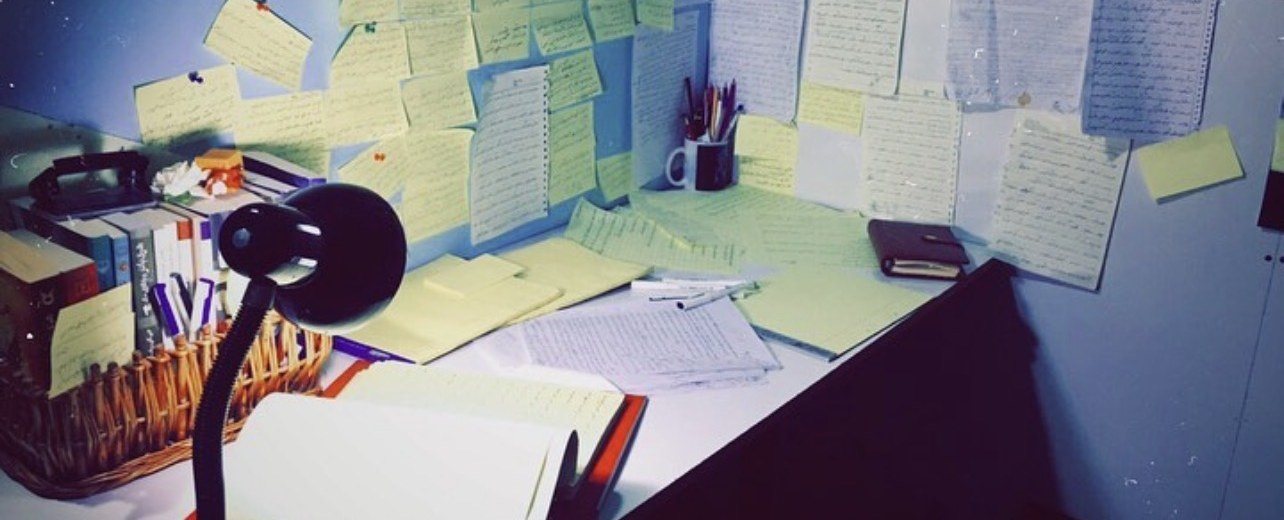 A writing desk under a lamp, the wall above it covered with handwritten pages and notes in Persian